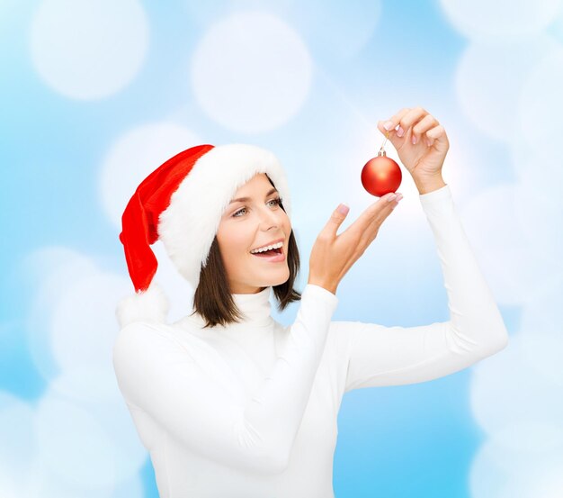 winter, people, christmas and happiness concept - woman in santa helper hat with christmas tree decoration ball over blue lights background