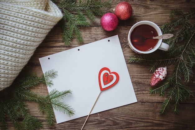 Зимний рисунок: чашка чая, еловые ветки, елочные игрушки, шарф и лист бумаги с сердечком на палочке на фактурной деревянной поверхности.