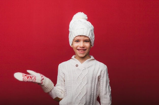 Winter portrait of a happy joyful childa boy in a knitted hat and sweater having fun on a red background the boy looks at his palms