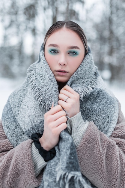 Winter portrait of a pretty young russian girl model in\
fashionable winter clothes with a vintage warm scarf, a fur coat\
and a sweater stands in the forest with snow