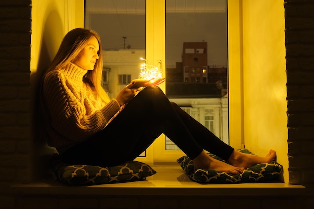 Winter portrait of young beautiful woman sitting near window on windowsill, night festive city background, sparkling garland lights in hands of girl