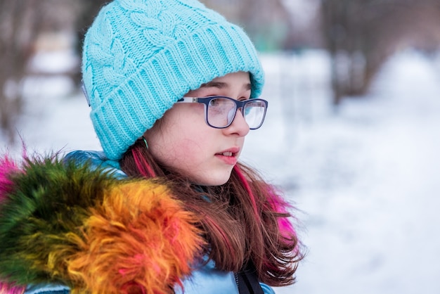 彼女の暖かい服を着た若い女の子の冬の肖像画。雪の降る天気で青い帽子をかぶった10代の少女。
