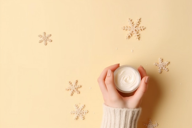 Winter season skin care cosmetics concept first person top view photo of woman's hands in knitted sweater small cream jar and snowflakes on isolated pastel beige background with empty space