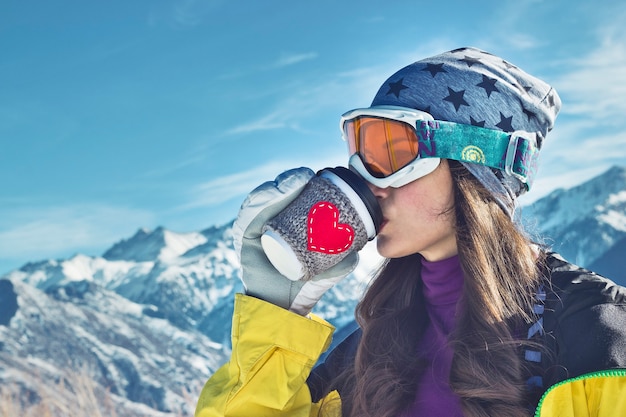 Девушка зимних видов спорта пьет из бумажного стаканчика с изображением сердца