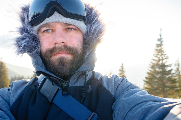 겨울, 스포츠 및 사람 개념 - 프리라이더 스노보더는 산봉우리의 가파른 경사면에 서서 스키 리조트의 눈 덮인 산을 배경으로 카메라나 스마트폰으로 셀카 초상화를 찍습니다.
