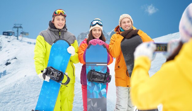 winter sport, technology, leisure, friendship and people concept - happy friends with snowboards and smartphone taking picture over snow and mountain background