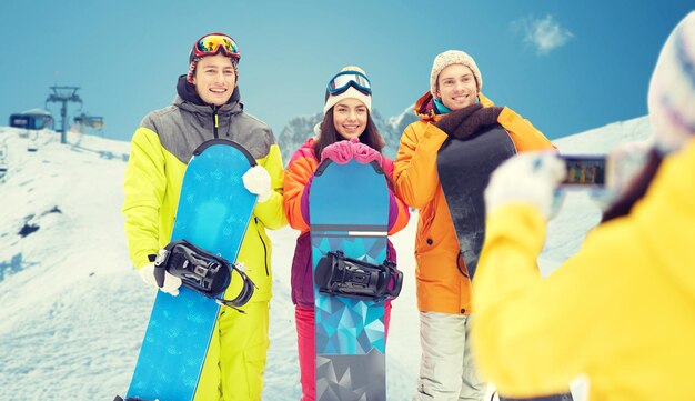 Winter sport, technology, leisure, friendship and people concept - happy friends with snowboards and smartphone taking picture over snow and mountain background