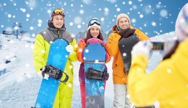 冬のスポーツ、テクノロジー、レジャー、友情、人のコンセプト – スノーボードとスマートフォンで雪と山の背景に写真を撮る幸せな友達