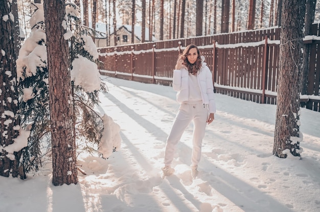 国の木造住宅の裏庭で白い暖かい上着ダウンジャケットの美しい若い女性の降雪の冬の日当たりの良い寒い肖像画