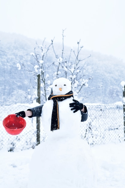 Winter teenager in a jacket and hat sculpts a snowman on a winter background with falling snow in frozen day concept