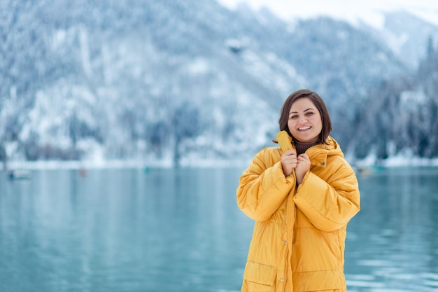 ヨーロッパを横断する冬の旅行。冬の高山湖のほとりに黄色いジャケットを着た美しい若い女性の肖像画。雪の高山湖の眺め。