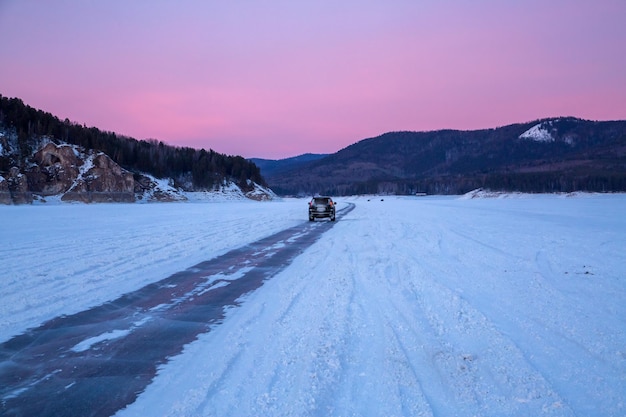 冬の旅 山と夕焼け空を背景に凍った川床に沿って凍った道を車で走る