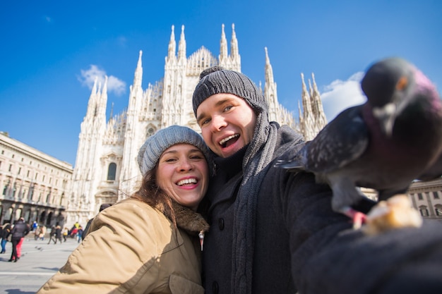 Winter travel and vacations concept - happy tourists taking a\
self portrait with funny pigeons in front of duomo cathedral in\
milan