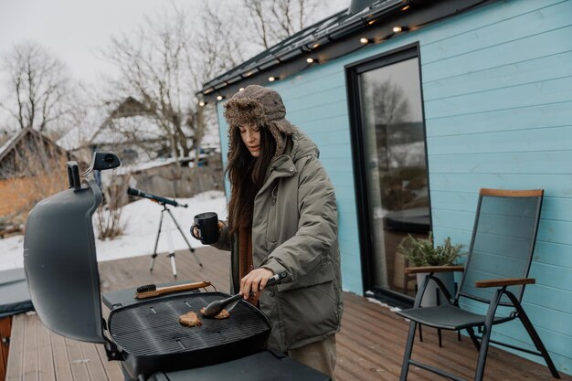 winter vacation in the backyard of a country tourist house A young stylish woman is grilling meat on a gas grill for her friends and family