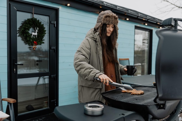 winter vacation in the backyard of a country tourist house A young stylish woman is grilling meat on a gas grill for her friends and family