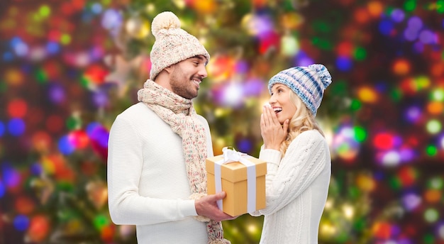 winter, vakantie, paar, kerst en mensen concept - lachende man en vrouw in hoeden en sjaal met geschenkdoos over lichten achtergrond