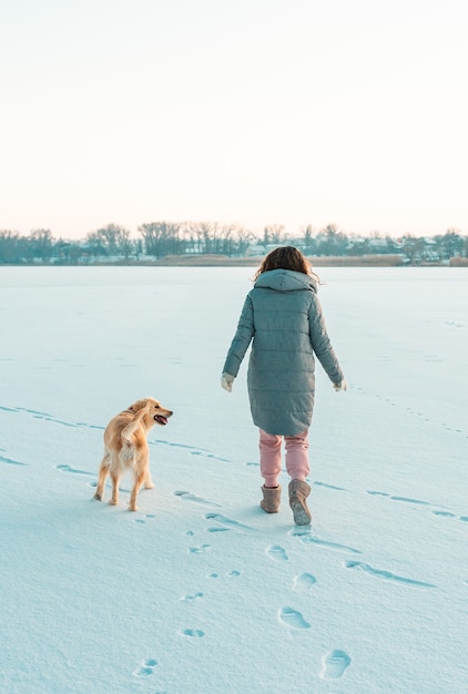 ゴールデン犬と雪の中で暖かい服を着ている若い美しいブルネットの女性の冬の散歩。雪の冬のコンセプト。