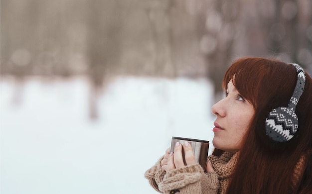 Winter. Woman with red hair wearing ear muffs. Girl drinking hot tea or coffee with iron insulated cup.