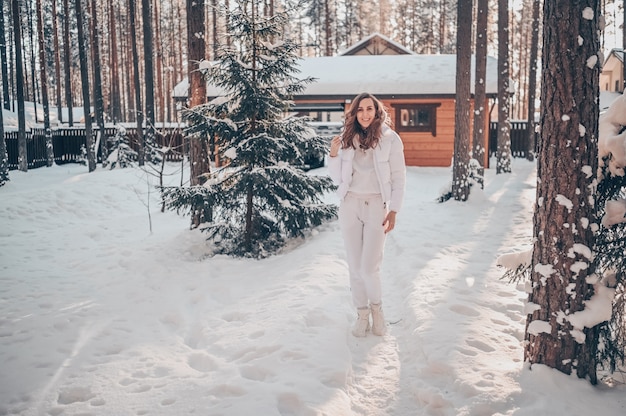 Winter zonnige koude portret in sneeuwval van mooie jonge vrouw in een witte warme uitloper donsjack in achtertuin van houten landhuis