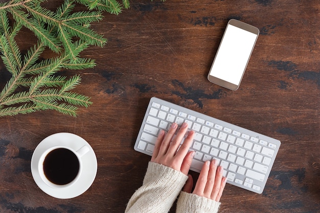 Wintergroene dennenboomtakken met kopje thee, mobiele telefoon en wit computertoetsenbord op houten ondergrond, plat leggen en van bovenaf bekijken