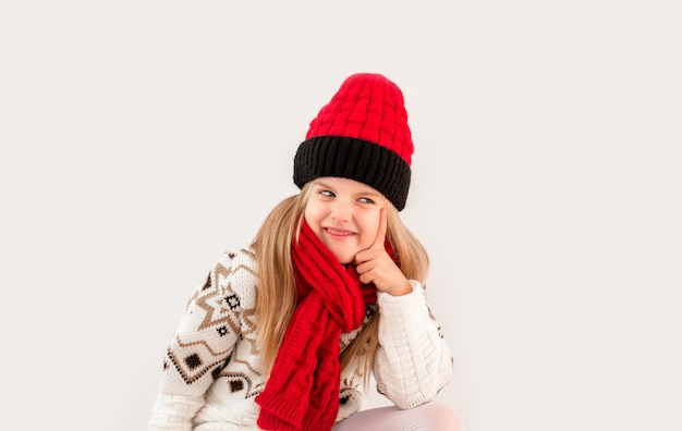 Winterkledij. Portret van een klein meisje met krullend haar in een gebreide witte wintermuts. klein blond meisje in witte gebreide muts en trui glimlachend lichte achtergrond isolate