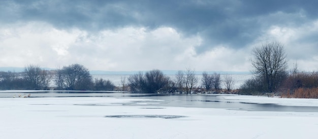 Winterlandschap met overdekte ijs- en sneeuwrivier, bomen aan de rivieroever en bewolkte hemel