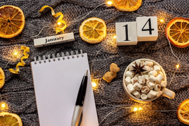 Winterse samenstelling. houten kalender 14 januari kopje cacao met marshmallow, lege open kladblok met pen, gedroogde sinaasappelen, lichte slinger op grijze gebreide achtergrond. bovenaanzicht platliggend mockup