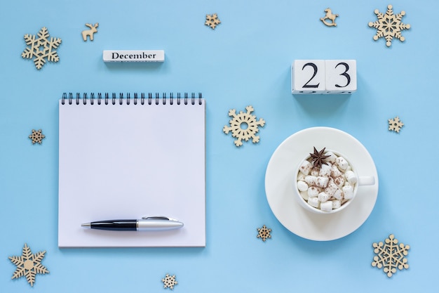 Winterse samenstelling. Houten kalender 23 December Kopje cacao met marshmallow en steranijs, lege open Kladblok met pen en sneeuwvlok op blauwe achtergrond. Bovenaanzicht Platliggend Mockup Concept