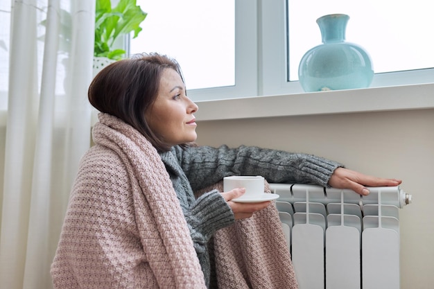 Winterseizoenvrouw die zich opwarmt in de buurt van de verwarmingsradiator van het huis