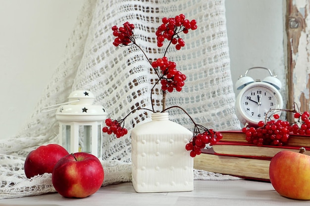 Winterstilleven een boeket viburnumtakken met rode bessen in een vierkante vaas rode appels bij een stapel boeken een klok tegen een gebreid gordijn