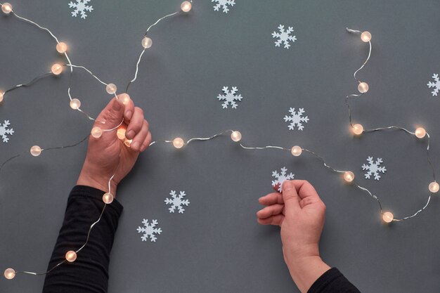 Wintervakantie feestelijke samenstelling. Hand met keramische fir tree decoratie. Nieuwjaar of Kerstmis plat lag. Handen houden lichte slinger en sneeuwvlok. Kerstmis bovenaanzicht op grijs papier achtergrond.