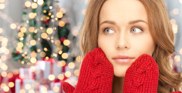 wintervakantie, kerstmis en mensenconcept - gelukkige jonge vrouw in rode wanten die haar gezicht aanraakt over de achtergrond van kerstboomverlichting