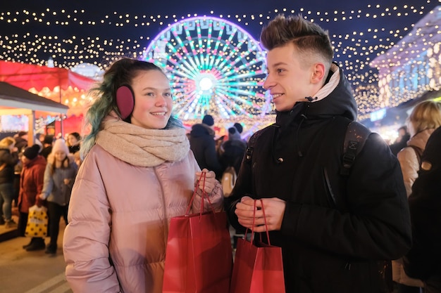 Winterwinkelen, kerstkortingen en geschenken, een paar tieners met boodschappentassen op de stadsmarkt, slingerlichten van de achtergrond van de avondstad