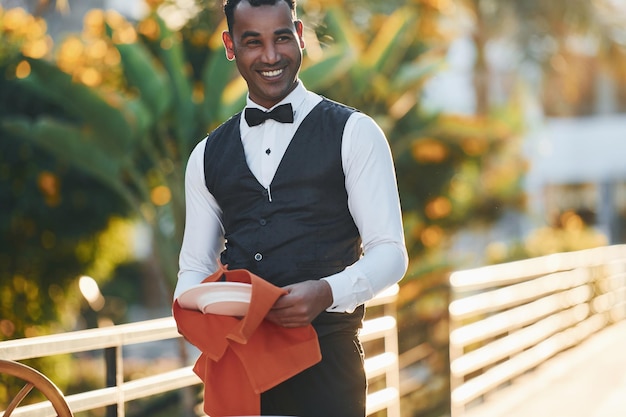 Wipes plates by red cloth Black waiter in formal clothes is at his work outdoors at sunny daytime