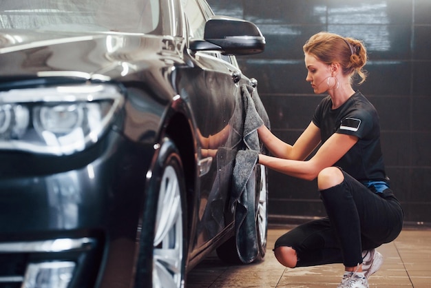 Wipes surface by using towel Modern black automobile get cleaned by woman inside of car wash station