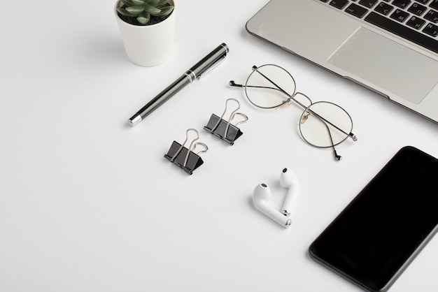 Беспроводные наушники, очки, ручка, зажимы, клавиатура мобильного телефона и ноутбука на белом столе, который является рабочим местом сотрудника