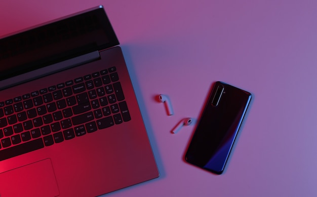 Wireless headphones and smartphone with laptop in neon red-blue gradient light. Top view