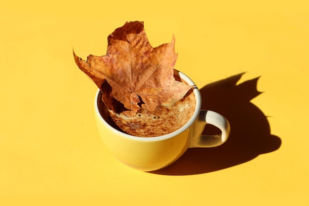 Пожелания уютных осенних дней желтая чашка кофе в ней сухой кленовый лист на желтом фоне тень от предметов вид сверху место для текста
