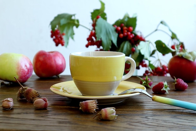 Wishes for a warm autumn morning a yellow cup of tea a teaspoon nuts on a wooden table against the background of viburnum branches and ripe apples side view space for text