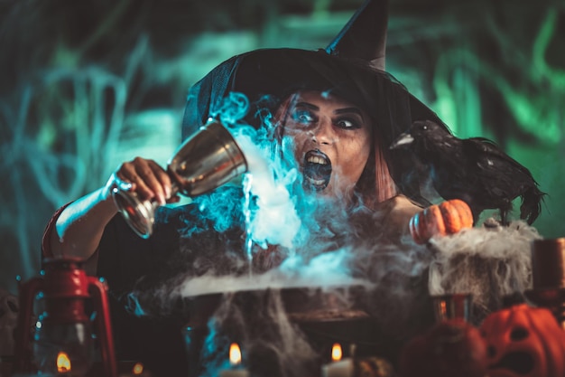 A witch in foggy surrounding holds a goblet above boiling water for a cooking poison soup.