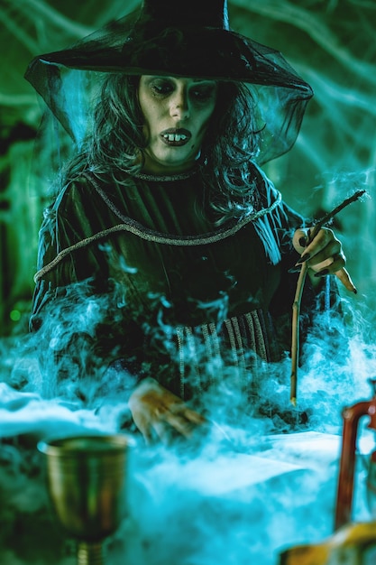 Photo witch with awfully face in creepy smoky green surroundings full of cobweb is reading recipe of magic drink. halloween concept.