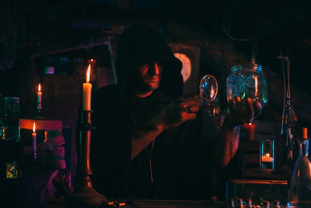 Witcher alchemist makes a secret potion in gloomy laboratory workshop. Dark magician sorcerer in Halloween costume at table