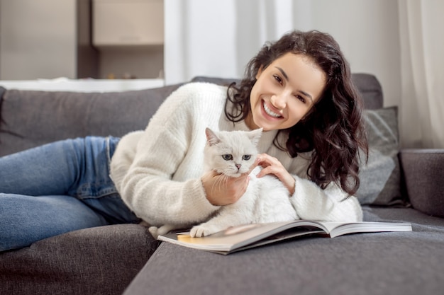 With a cat. Cute woman laying on the sofa and caressing her cute white cat