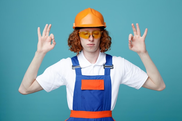 With close deyes showing okay gesture young builder man in uniform isolated on blue background