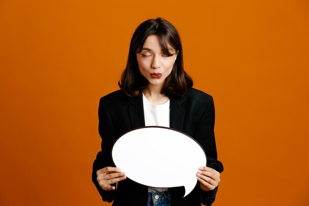 With closed eyes holding speech bubble young beautiful female wearing black jacket isolated on orange background