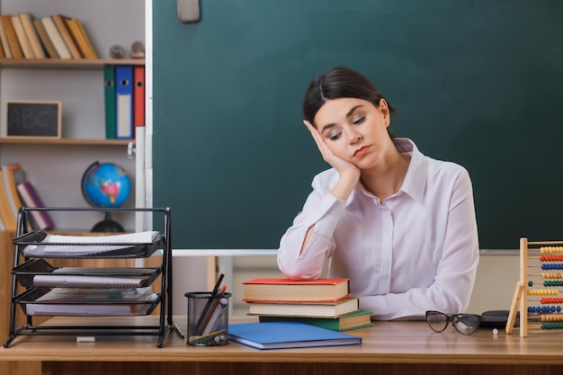 with closed eyes putting hand on cheek young female teacher sitting at desk with school tools in classroom