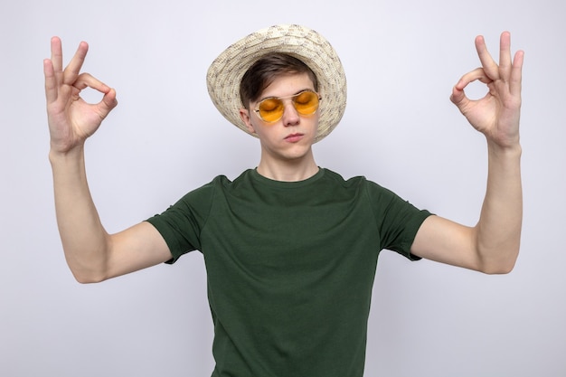 With closed eyes showing meditation young handsome guy wearing glasses with hat 