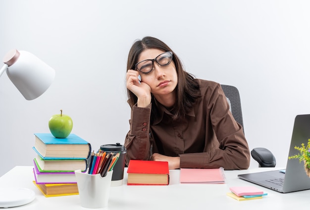 With closed eyes tired young school woman wearing glasses sits at table with school tools putting hand on cheek 