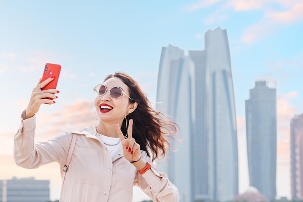 With a confident smile and a smartphone in hand a girl captures a beautiful moment in front of the towering skyscrapers of Abu Dhabi