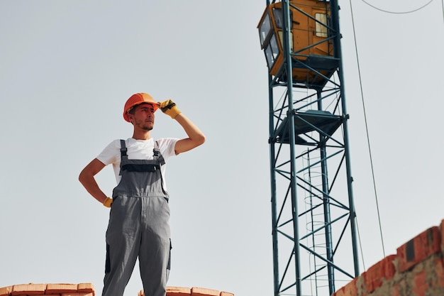 With crane at background Young construction worker in uniform is busy at the unfinished building